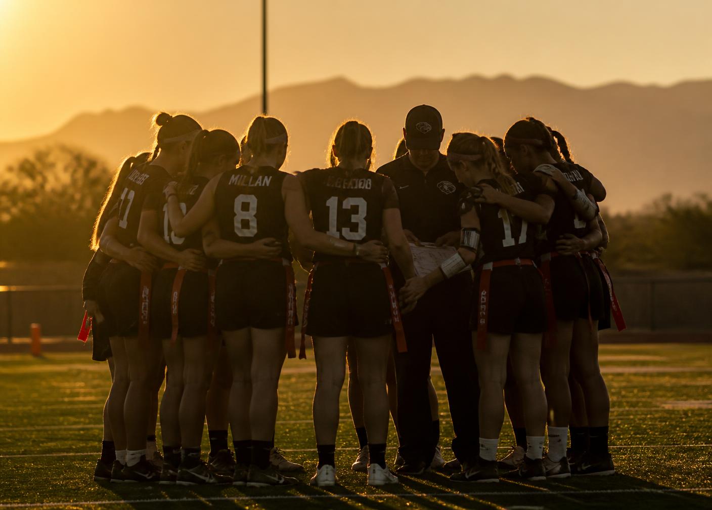 Girls flag football team huddled with a coach at sunset